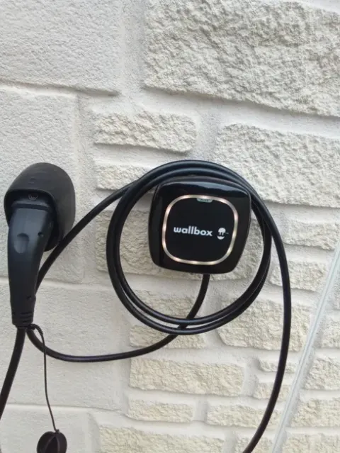 Borne de recharge murale noire avec son câble Type 2 lové, sur un mur en pierre